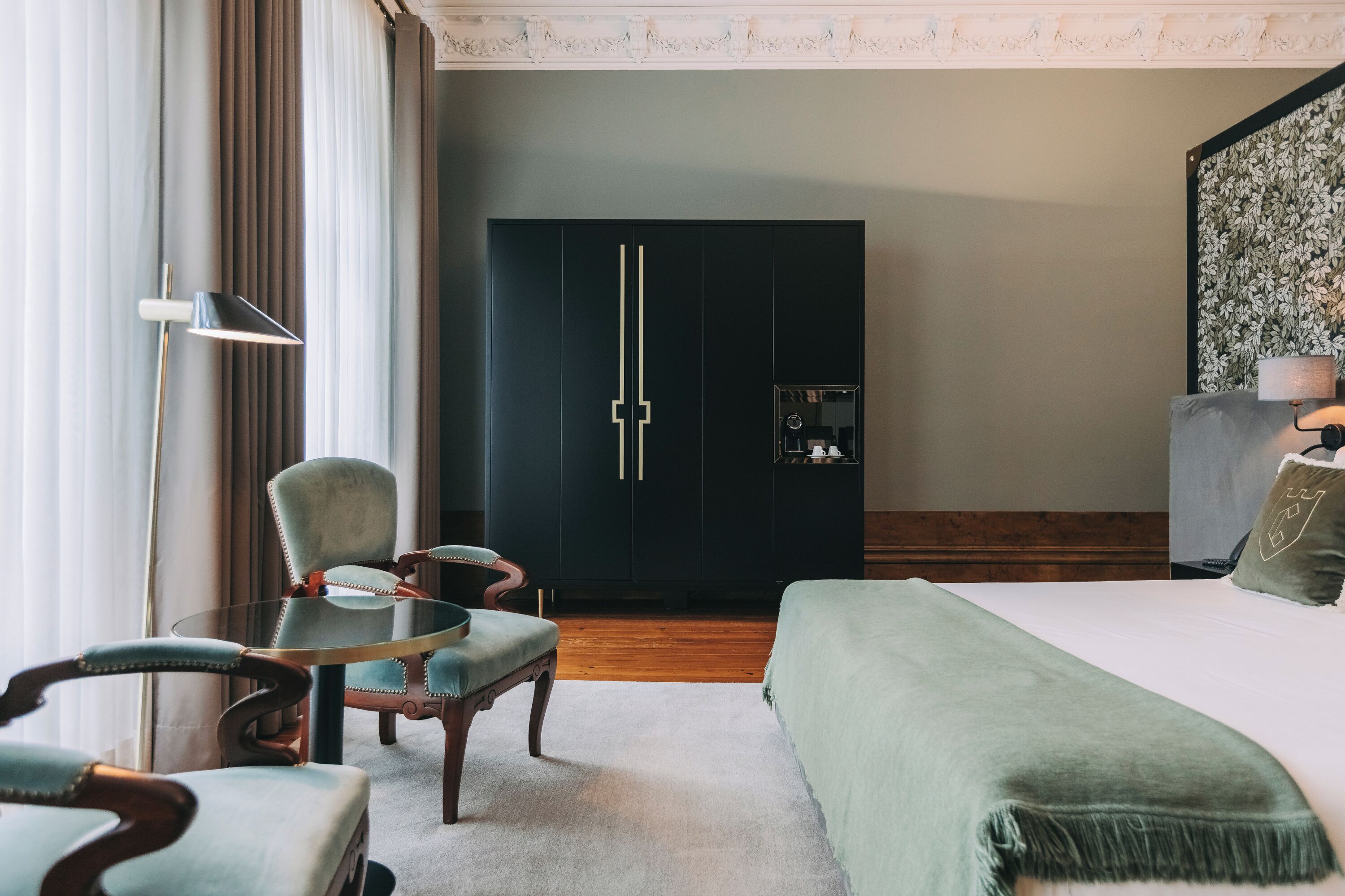 A hotel room with a bed, a desk with a chair, a wardrobe, and a lamp.