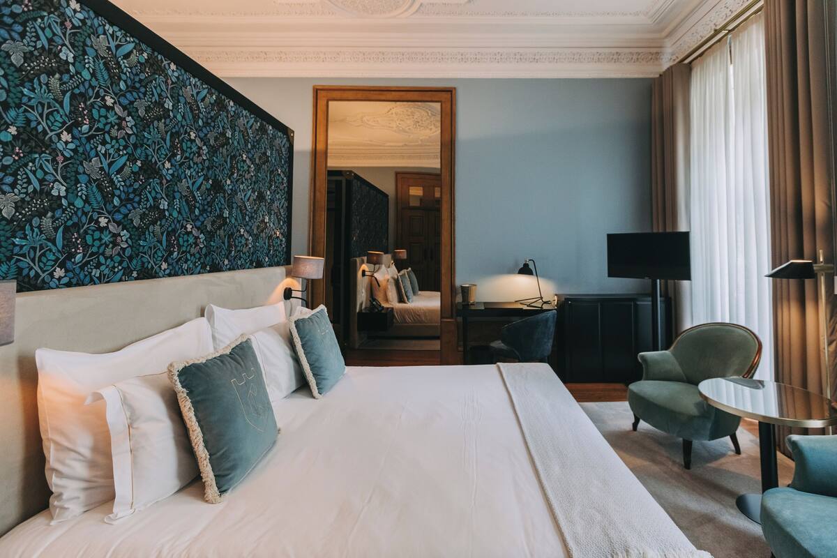 A hotel room with a large bed, a desk, a chair, a TV, and a large window with curtains.