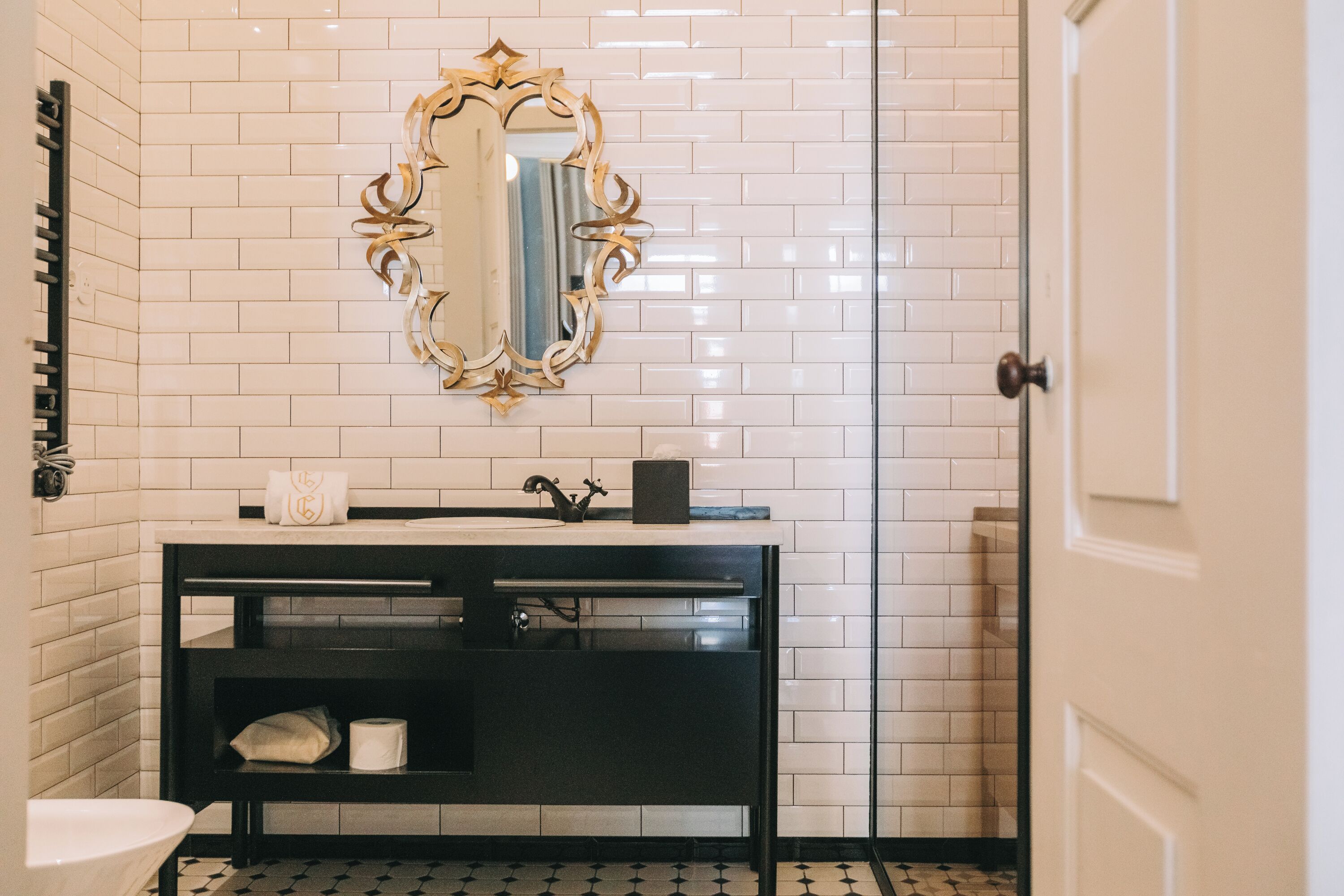 Een badkamer met witte betegelde wanden, een zwart wastafelmeubel, een spiegel met gouden lijst en een wit toilet.