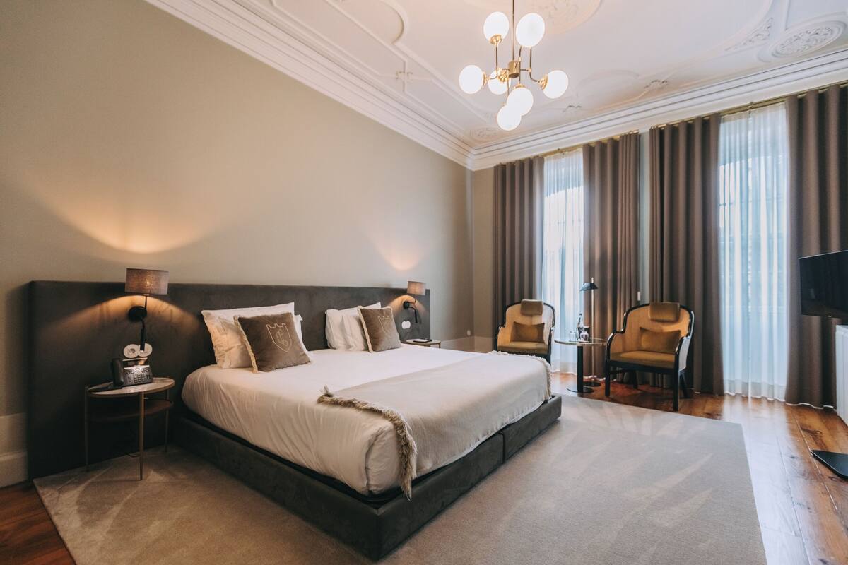 A spacious bedroom with a large bed, a nightstand, a chair, a TV, and a chandelier.