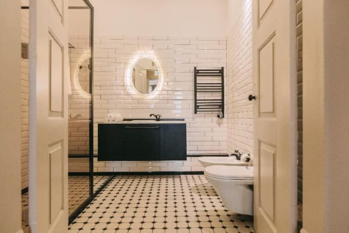 A bathroom with a black and white tiled floor, a black vanity, a white toilet, and a mirror with a circular light.