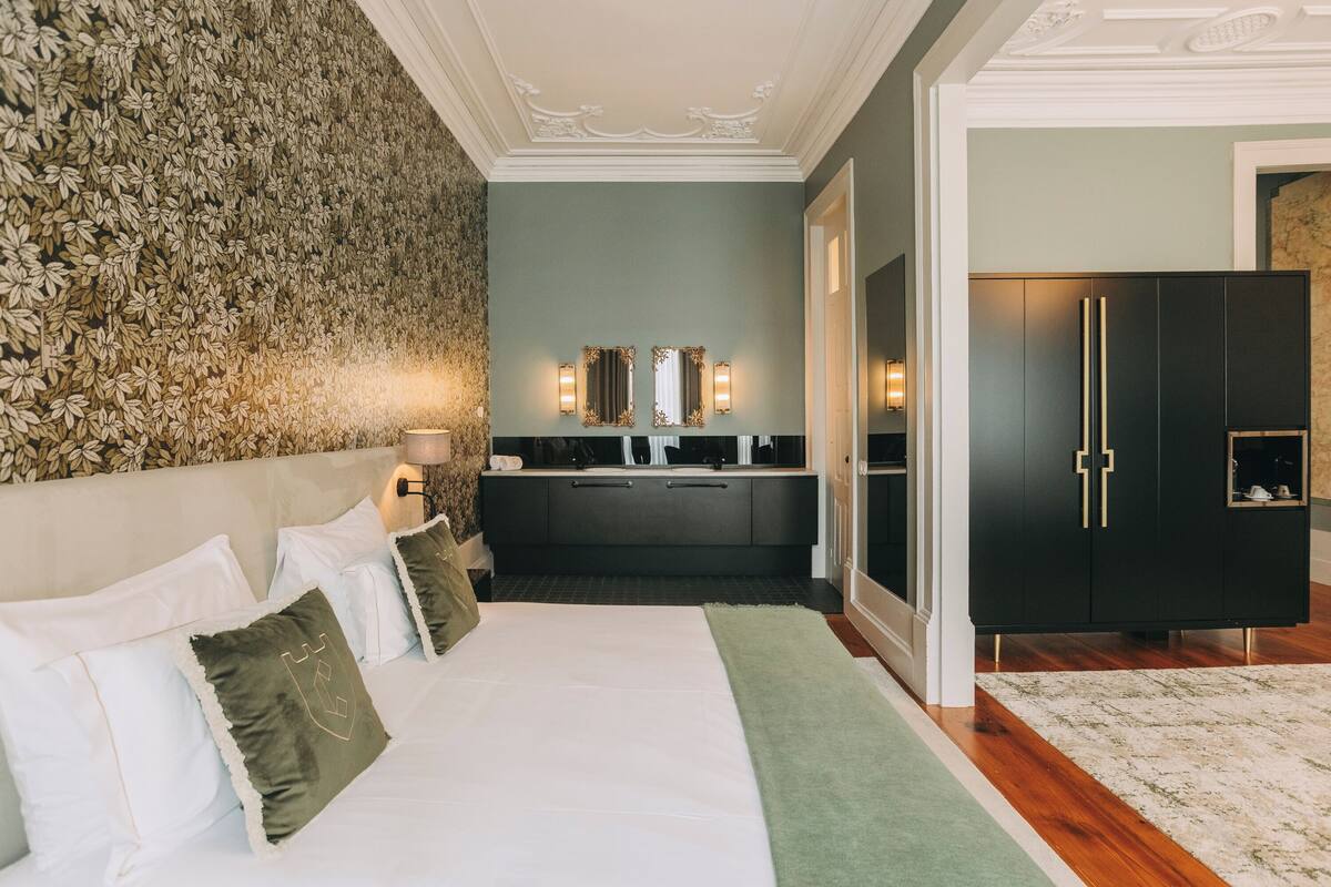 A bedroom with a bed, pillows, a nightstand, a mirror, a dresser, and a wall with a floral pattern.