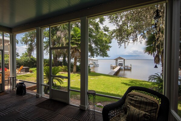 A covered outdoor area with a view of a lake, a dock, and a grassy area with chairs and a table.