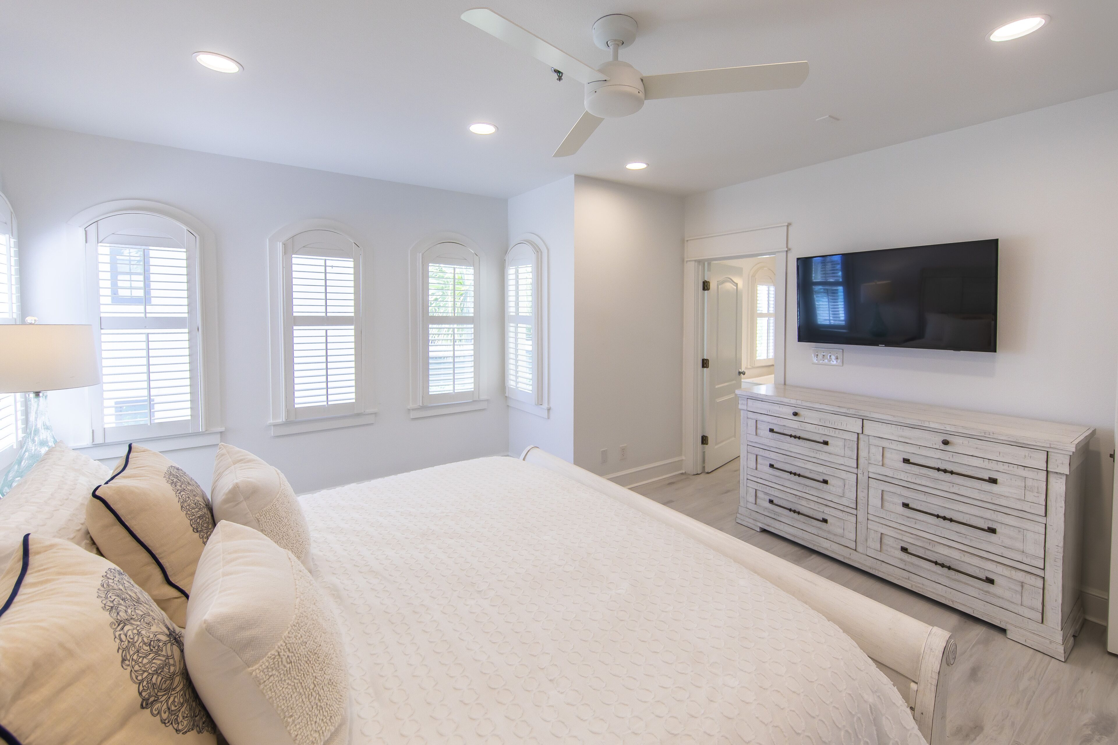 A bedroom with a large bed, a ceiling fan, a TV mounted on the wall, and a dresser with drawers.