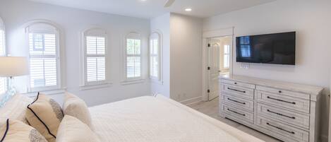 A bedroom with a large bed, a ceiling fan, a TV mounted on the wall, and a dresser with drawers.