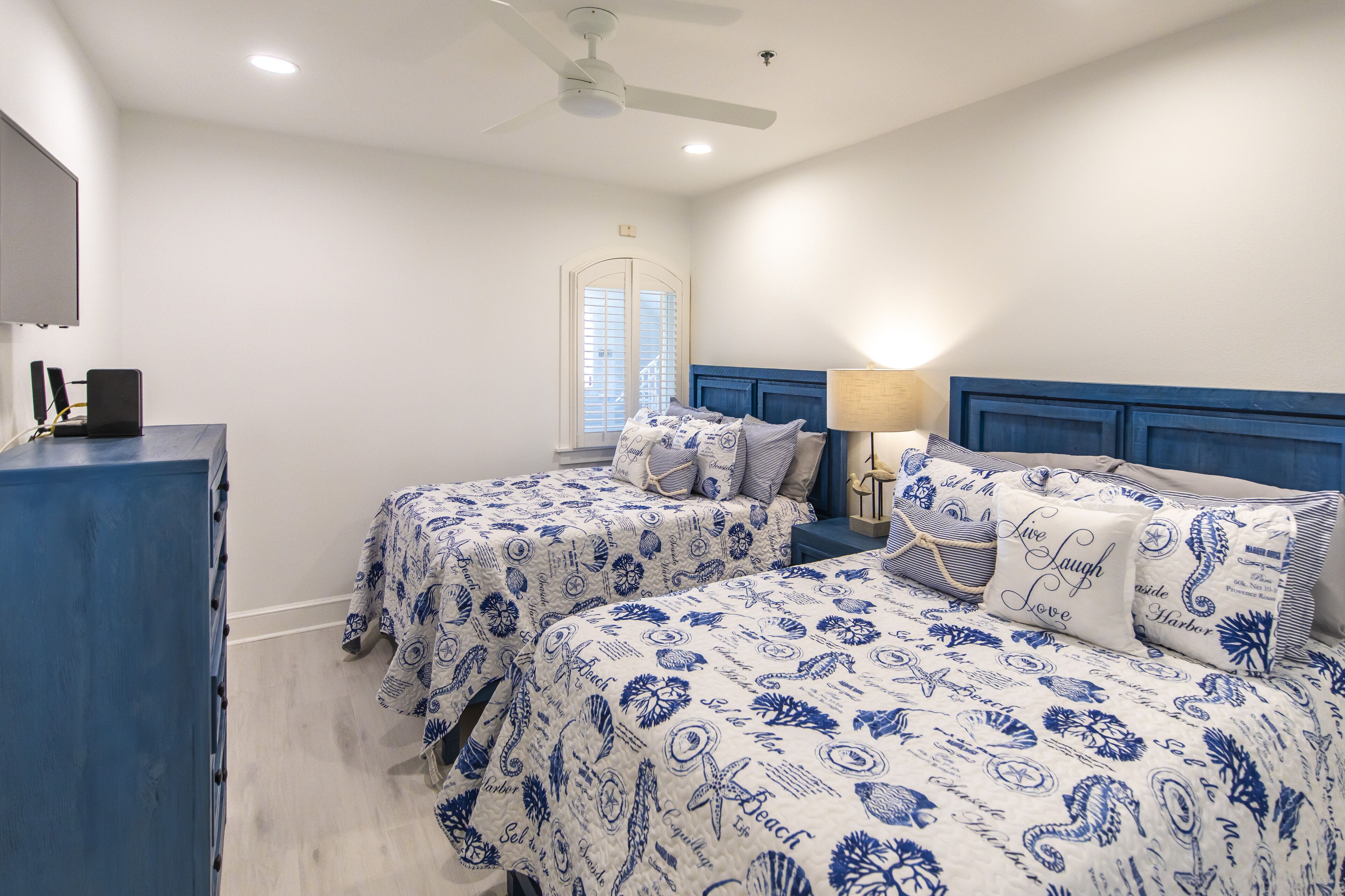 A bedroom with two beds, a dresser, a ceiling fan, and a window with shutters.