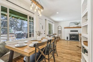 A dining area with a wooden table set for four, surrounded by chairs, a fireplace, and large windows offering a view of trees.