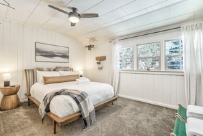 Un dormitorio con una cama, ventilador de techo, ventana con cortinas e imagen colgada en la pared.