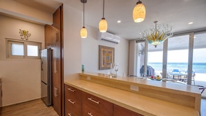 A modern kitchen with wooden cabinets, a stainless steel refrigerator, pendant lights, and a view of the ocean through large windows. - Beach-Level Escape Featuring a New Private Plunge Pool and Ocean Views! (Akumal)
