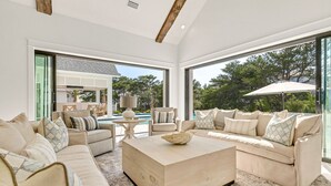 A modern living room with a beige sofa set, a wooden coffee table, and a large window overlooking a pool and greenery.
