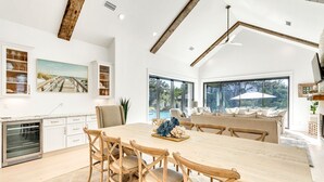 Un comedor espacioso con una mesa grande y sillas de madera, una cocina con armarios blancos y vistas a una piscina a través de amplios ventanales.