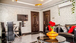 A modern living room with a marble floor, a black leather sofa, a glass coffee table with a vase of flowers, and a flat-screen TV mounted on the wall. - Tobby Royal Suites (Lagos)