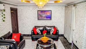 A living room with a black leather sofa, a glass coffee table with a vase of flowers, a chandelier, and a framed painting on the wall. - Tobby Royal Suites (Lagos)