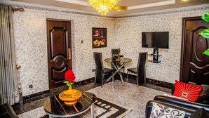 A well-lit room with a chandelier, a dining area with a round table and chairs, a television mounted on the wall, and a door with a decorative handle. - Tobby Royal Suites (Lagos)