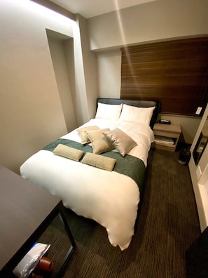 Una cama bien hecha con almohadas y una manta, una mesita de noche con un box de pañuelos, y una ventana con persianas.