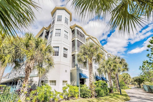 A multi-story building with a corner tower, surrounded by palm trees and greenery.
