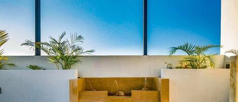 A modern outdoor spa area with a central water feature, surrounded by white walls and palm plants.