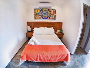 A hotel room with a bed, two nightstands, a wall-mounted air conditioner, and a colorful artwork above the bed.