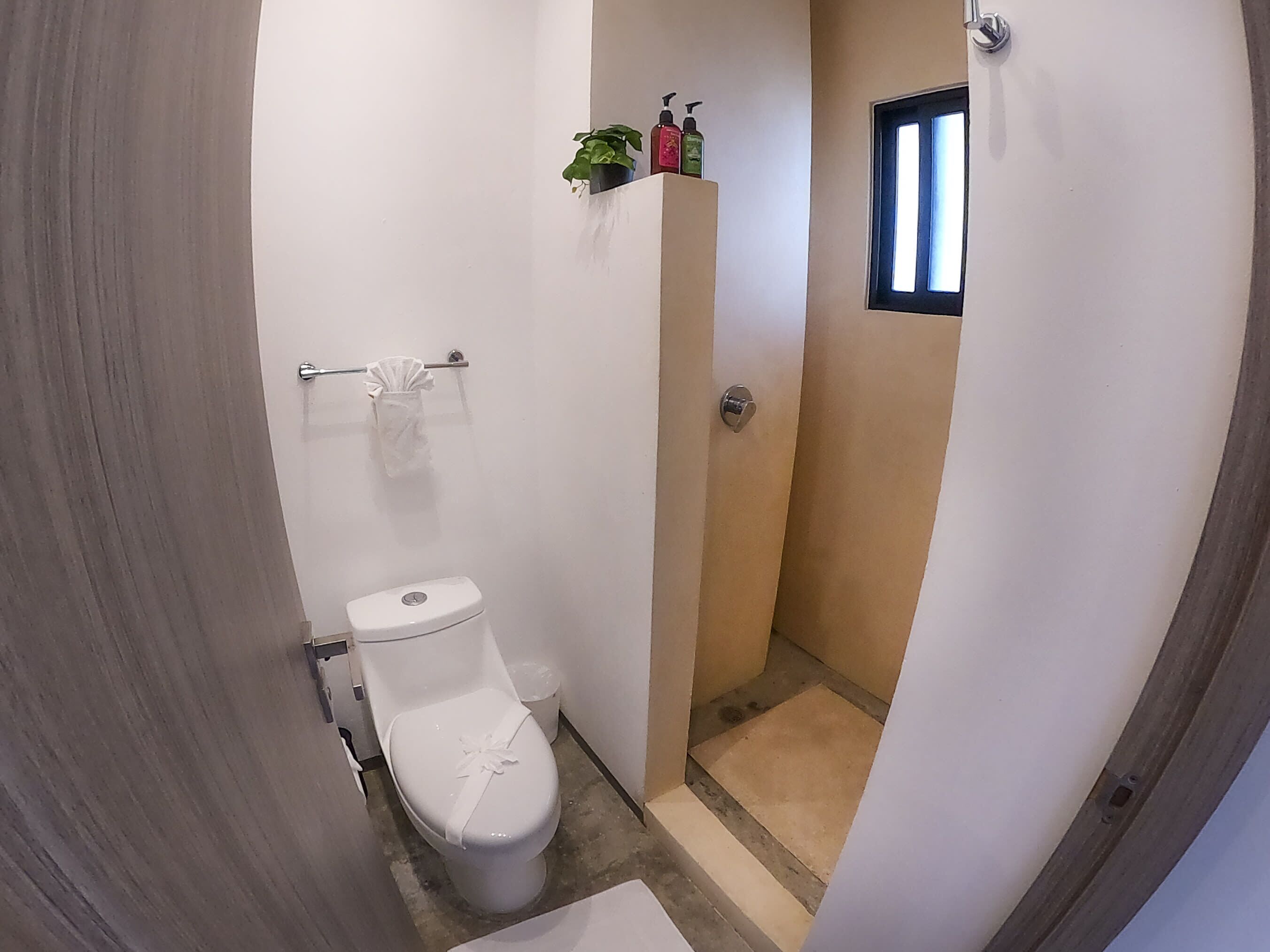 A small, narrow bathroom with a toilet, a small shelf with toiletries, and a window.