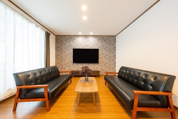Un salon moderne avec deux canapés en cuir noir, une table basse en bois et un téléviseur à écran plat fixé à un mur dont le motif rappelle celui d’un mur de briques.