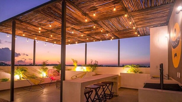 A rooftop terrace with a thatched roof, string lights, seating, and a view of the cityscape at dusk. - Stylish Condo - Pool + Rooftop - Walk to 5th Ave (Playa del Carmen)
