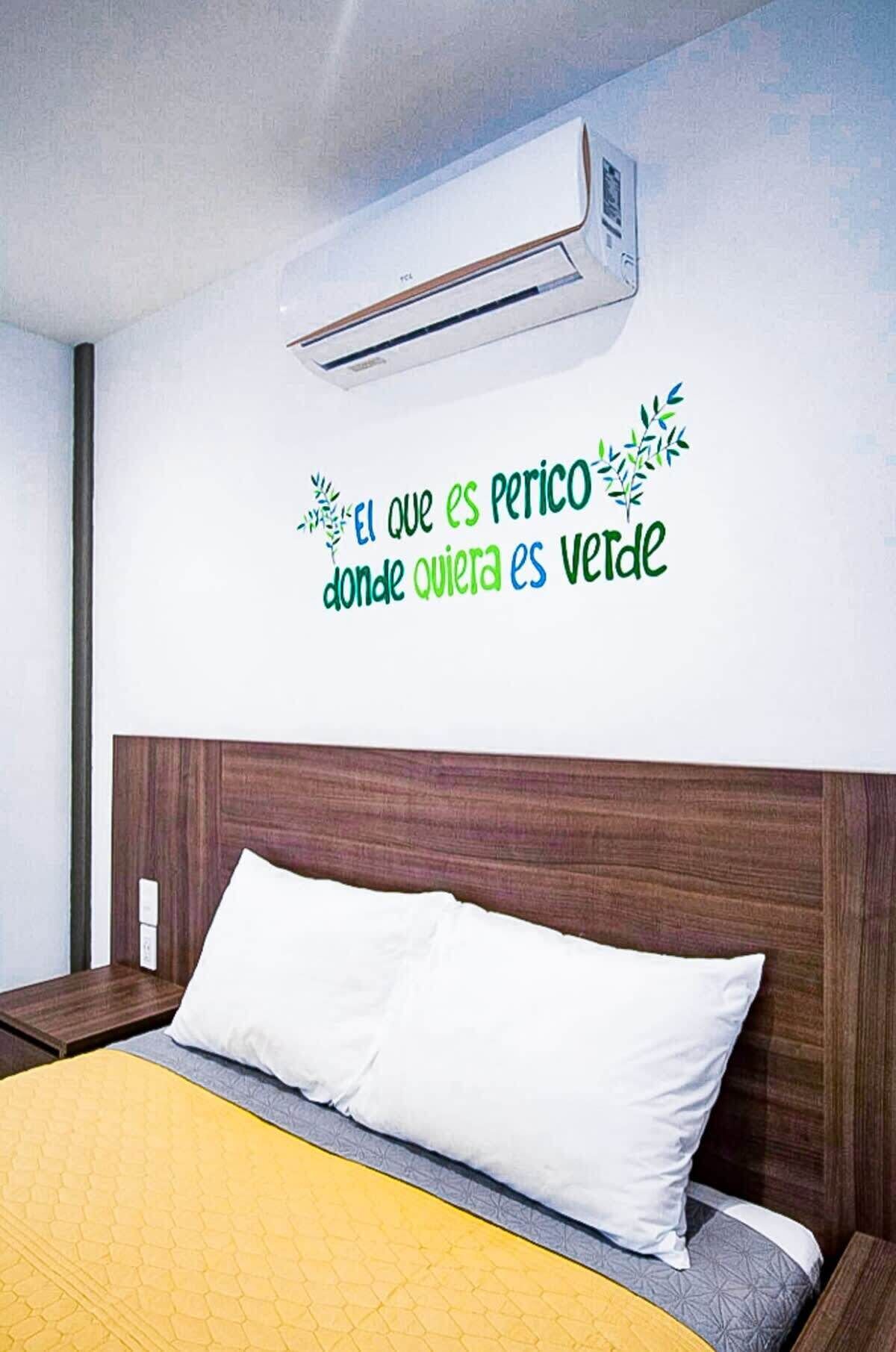 A hotel room with a bed, a wall-mounted air conditioner, and a decorative wall decal with text and leaf graphics.