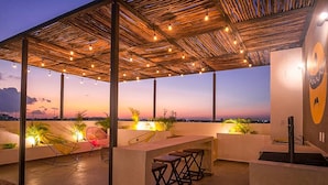 Uma área de estar ao ar livre com terraço coberto por palha, luzes de corda e banquetas de bar.