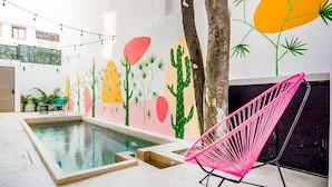 Uma área de piscina com um mural de cactos e palmeiras, uma cadeira rosa e luzes de cordão.
