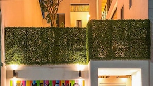 Entrada de hotel com um mural colorido, uma porta de madeira fechada e uma sebe verde.
