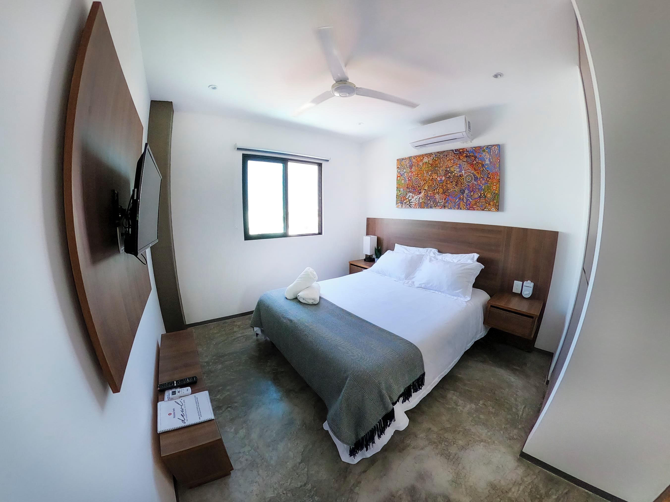 Hotelkamer met een bed, bureau, stoel, televisie, plafondventilator, airconditioning en een schilderij aan de muur.