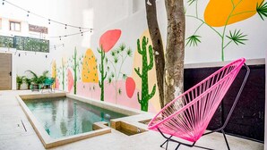 Zona de piscina amb un mural de cactus i palmeres, una cadira rosa i llums de cadena.