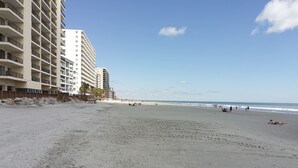 Ein Strand mit einem großen Gebäude links, blauem Himmel und Fußabdrücken im Sand.