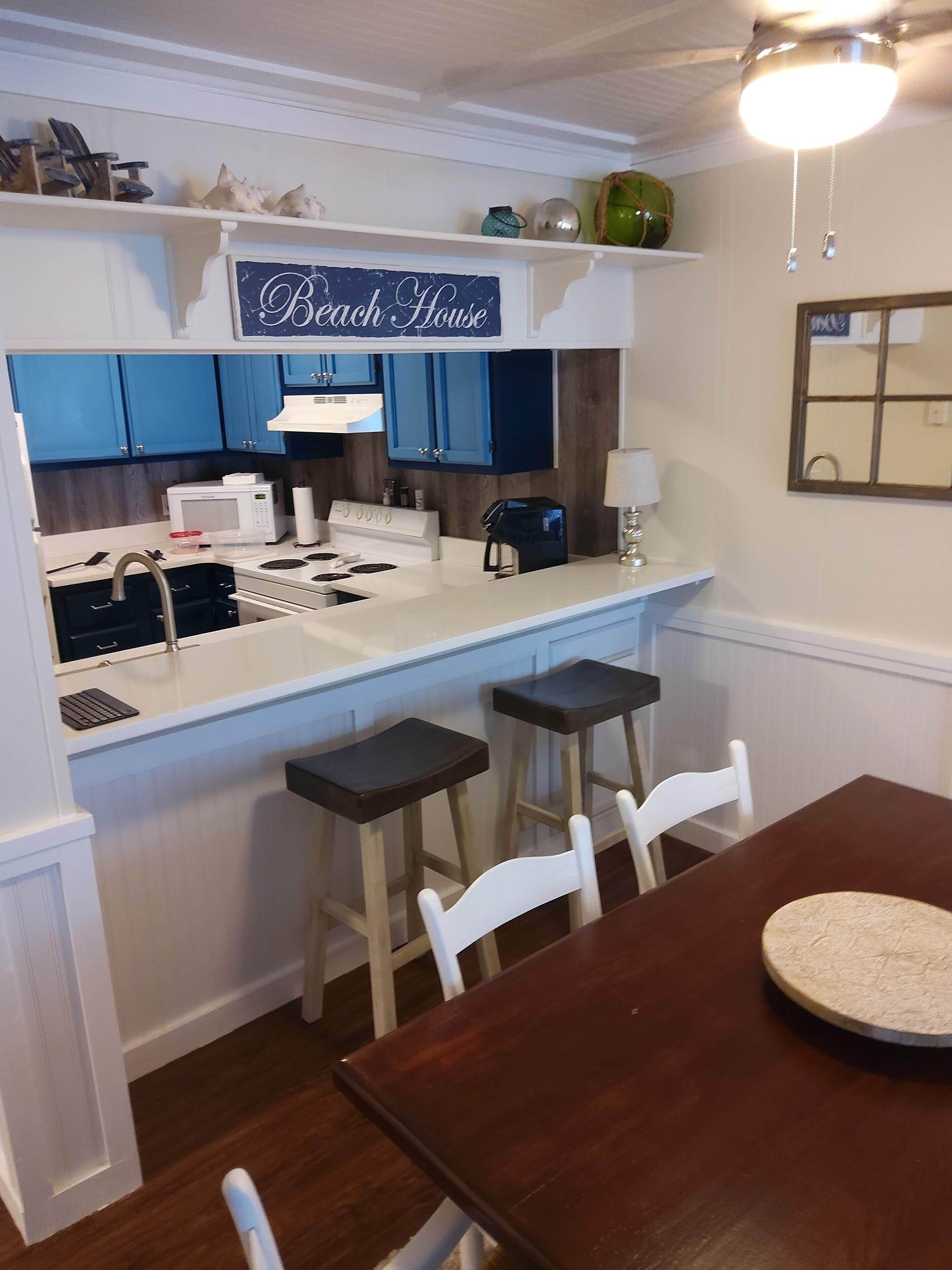 A compact kitchen with a microwave, stove, and coffee maker. There are two bar stools and a dining table with chairs. A sign above the counter reads "Beach House."