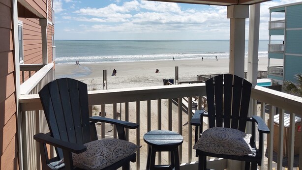 Ein Balkon mit zwei Adirondack-Stühlen und einem kleinen Tisch, der Blick auf einen Strand mit Menschen und einem Hund bietet.