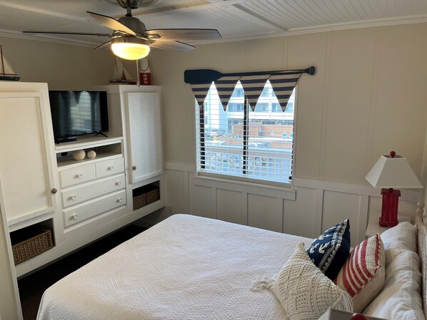 Un dormitorio con cama, televisión, ventilador de techo, ventana con cortinas a rayas y una lámpara roja.