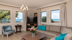 A living room with a sofa, armchair, coffee table, TV, bookshelf, and a view of the sea. - Villa Elizabeth   Seafront Classic Villa! (Zakynthos)