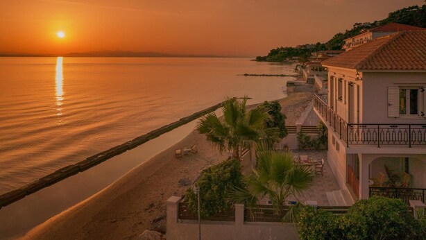 A coastal building with a balcony overlooking a beach, a pier extending into the water, and a sunset in the background. - Villa Elizabeth   Seafront Classic Villa! (Zakynthos)