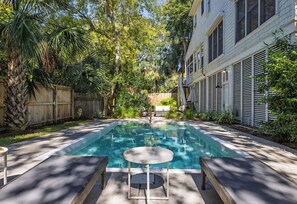 Una piscina rectangular con una mesa y sillas en primer plano, rodeada de abundante vegetación y una valla de madera.