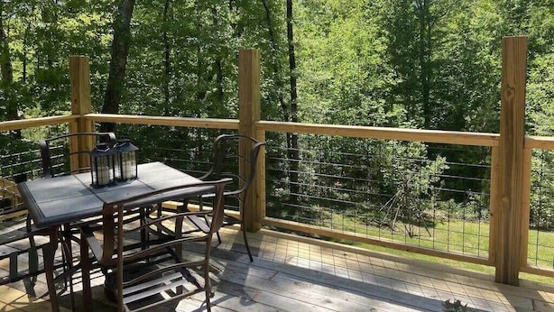 Une terrasse en bois, agrémentée d’une table et de chaises, entourée d’une balustrade en bois et d’une clôture en treillis métallique, et offrant une vue sur une zone boisée.