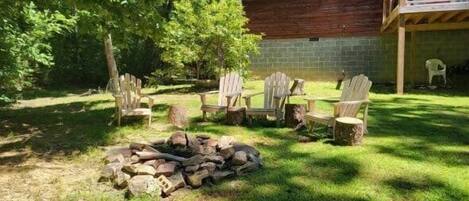 A wooden cabin with a covered porch, a fire pit, and Adirondack chairs in a grassy yard.