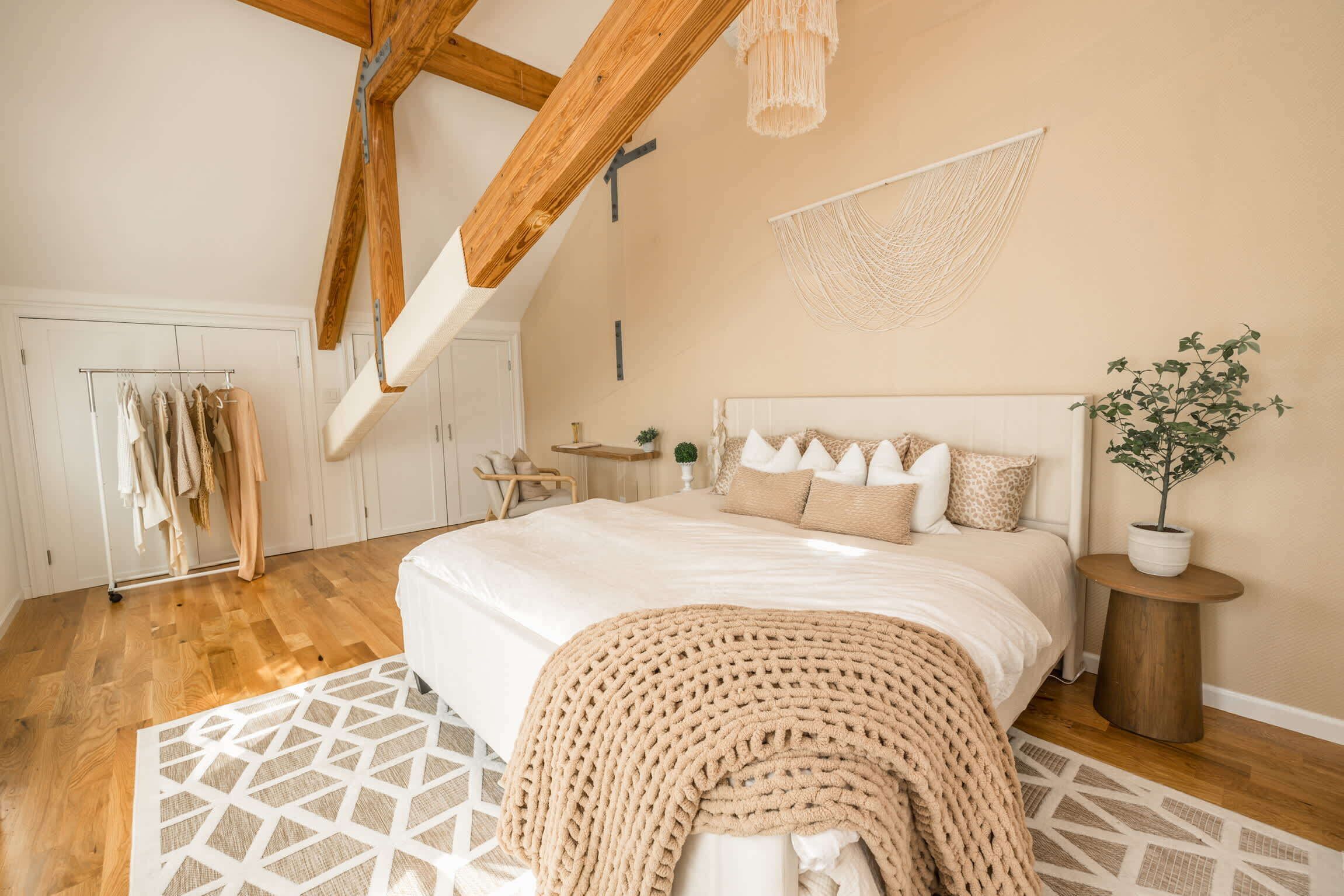 A bedroom with a large bed, wooden beams, a hanging macrame, a small round table, and a potted plant.