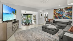 Ein modernes Wohnzimmer mit großem Flachbildfernseher, grauen Sofas, einem Essbereich mit Stühlen und einem farbenfrohen abstrakten Gemälde an der Wand.