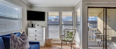 A modern living room with a flat-screen TV, a blue sofa, a wicker chair, and a balcony with outdoor furniture.