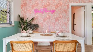 Ein Essbereich mit einem weißen Tisch, gedeckt für vier Personen, umgeben von Korbstühlen, vor einer Wand mit rosa floralem Wandpapier und einem Neon-Schild mit der Aufschrift 'Pink Flowers'.
