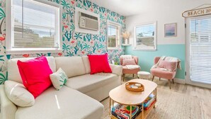 Ein Wohnzimmer mit einem weißen Sofa, rosa Sesseln, einem Couchtisch und einer Wand mit tropischem Muster.