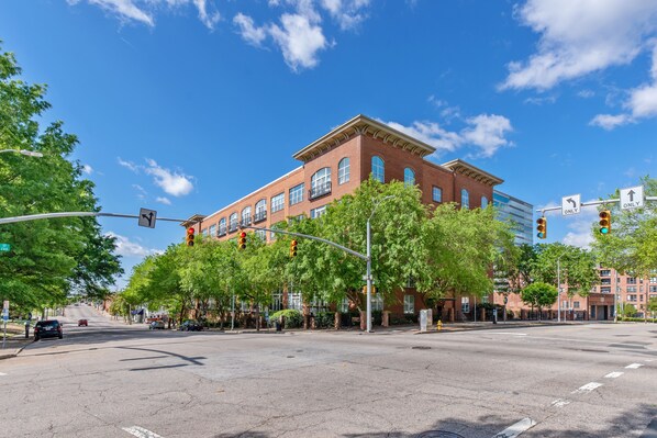 A street intersection with a large brick building, traffic lights, and trees. - One Bedroom Hideaway w/ free parking steps to Downtown (Raleigh)