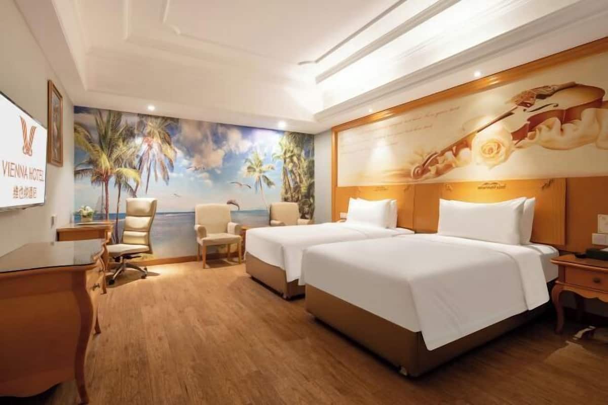 Ein Hotelzimmer mit zwei Betten, einem Schreibtisch, einem Stuhl, einem kleinen Tisch und einem Wandgemälde mit einer Strandansicht.