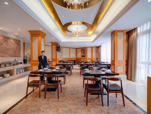 A spacious dining area with multiple tables and chairs, a central chandelier, and a buffet counter with food items.