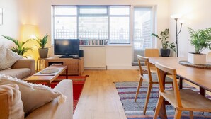 A modern living room with a sofa, a coffee table, a TV, a dining table with chairs, and large windows with blinds. - 3-Bedroom Duplex w/ Private Patio in Shoreditch - Pass the Keys (London)