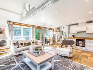A spacious living area with a sofa set, a coffee table, and a dining table. There is a kitchen area with modern appliances and a staircase leading to an upper level.